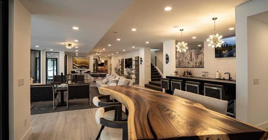 Indoor room with a large natural wood bar area with a living areas in back with pieces of art on the wall and a lot of hanging and ceiling lights