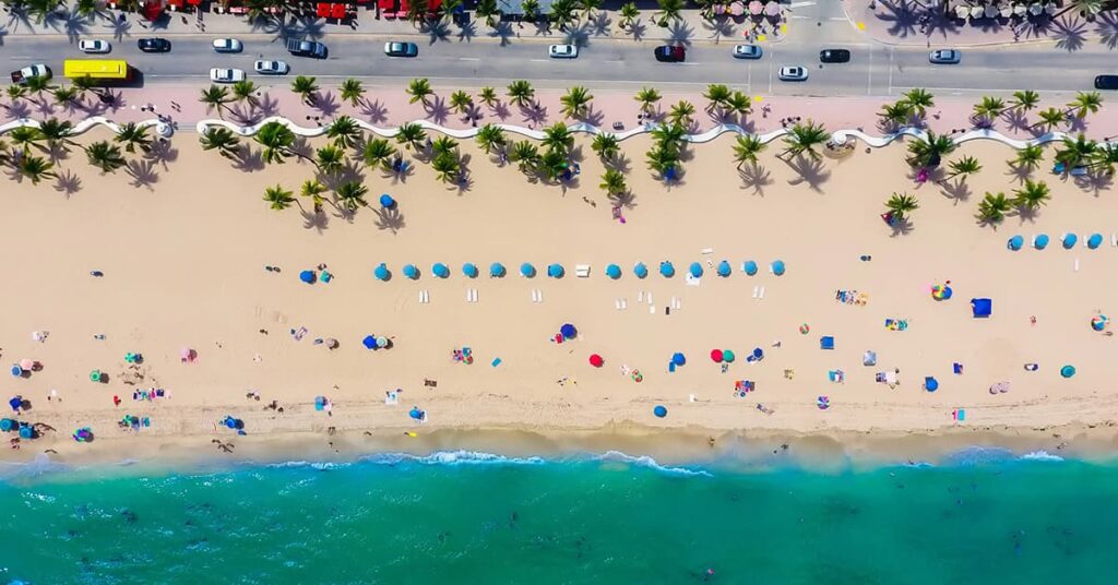 Overhead shot of a beach with sand, water and beach-goers in front of a row of palm trees and a road with cars on it