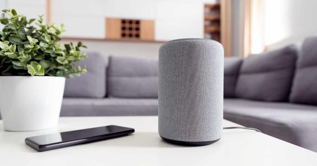 White table with gray Bluetooth speaker, smartphone, and plant on it with a gray sectional couch in the background