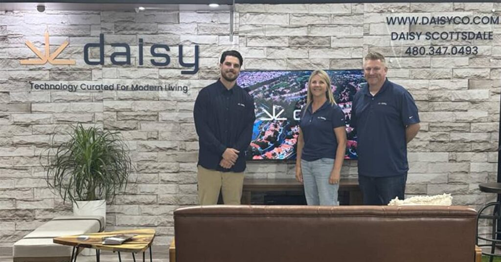 Three Daisy employees in front of a wall with the Daisy logo and Scottsdale information on it