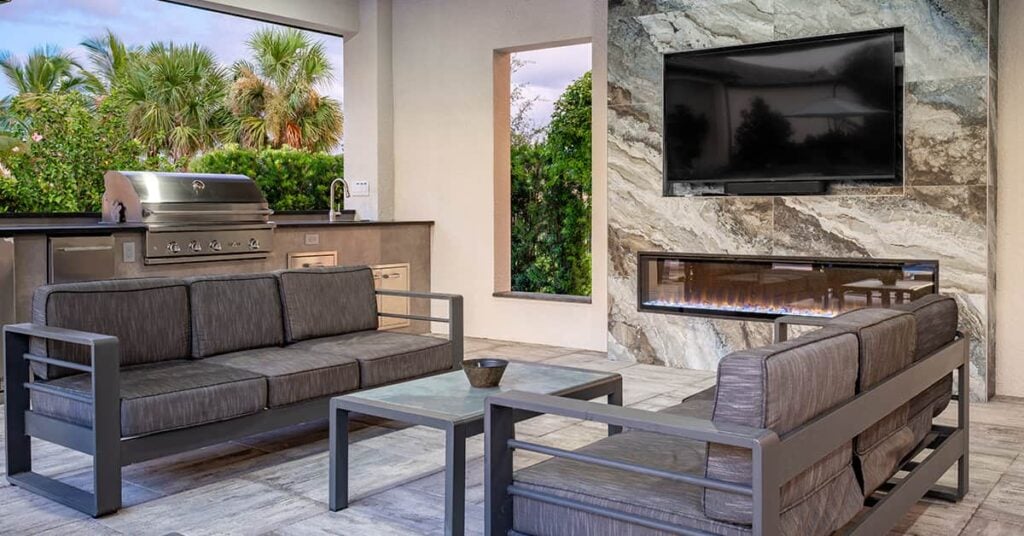 Enclosed open-air outdoor area with two grey couches a grilling station in back and a flat screen TV on a stone wall over a gas fireplace
