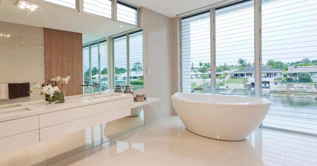 Luxury modern bathroom with stand-alone tub next to windows with blinds on it near a waterway with boats in it