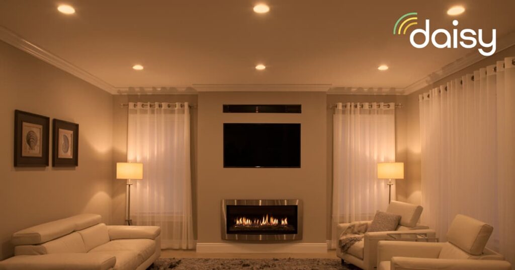 Living area in the evening with ambient lighting and a flat screen TV on the wall above a fireplace in between toe windows with curtains