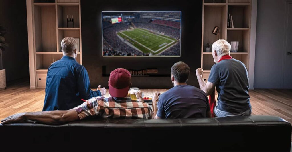 Rear view of four people sitting on a couch watching a football game on a flat screen TV