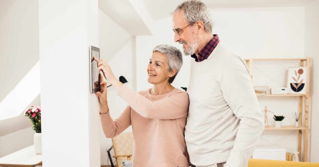 An elderly man and woman in a white room smiling while using a smart home control panel on a wall