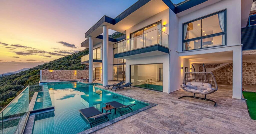 Modern home with two large columns in the evening with a pool area with lounge chairs to the left, a swinging chair to the right, and lights on in the house windows