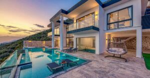 Modern home with two large columns in the evening with a pool area with lounge chairs to the left, a swinging chair to the right, and lights on in the house windows