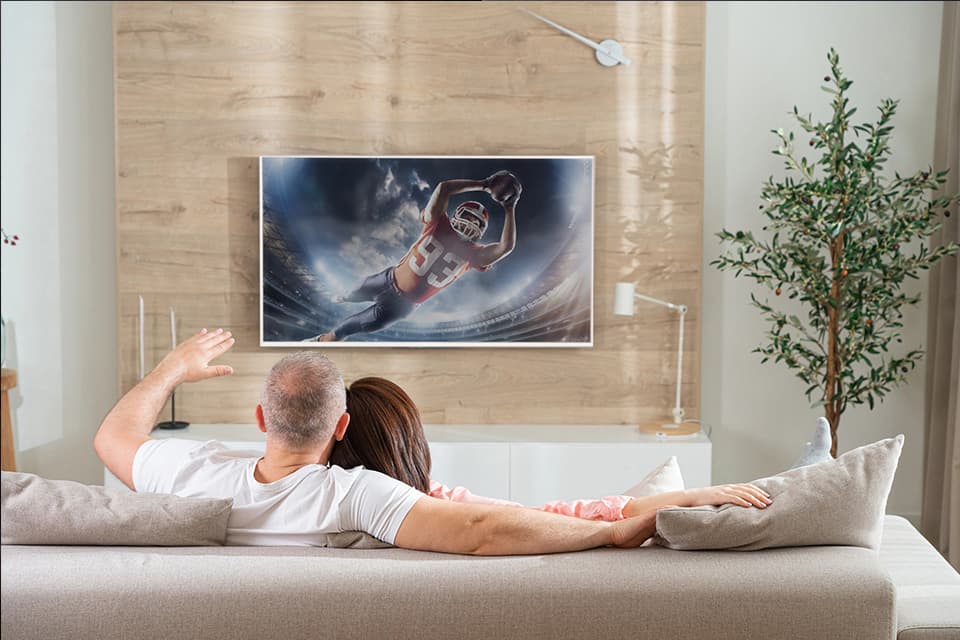Two people in a light colored room sitting on a white couch watching a flat screen TV on wall with football player on it