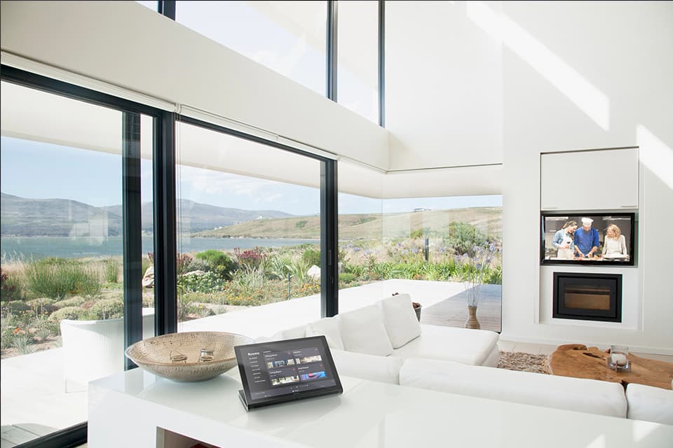 White indoor living area with large windows, a flat screen TV on the wall, and a white wrap-around couch with a table showing a tablet PC with security images on it