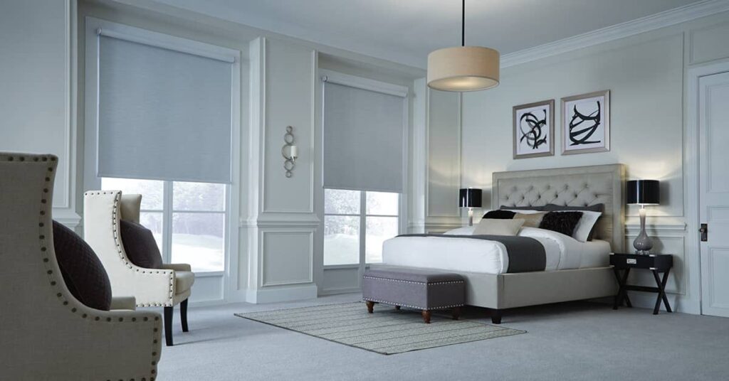 Light colored bedroom with two chairs on the left, two images on the wall behind the bed, and gray motorized shades on the windows