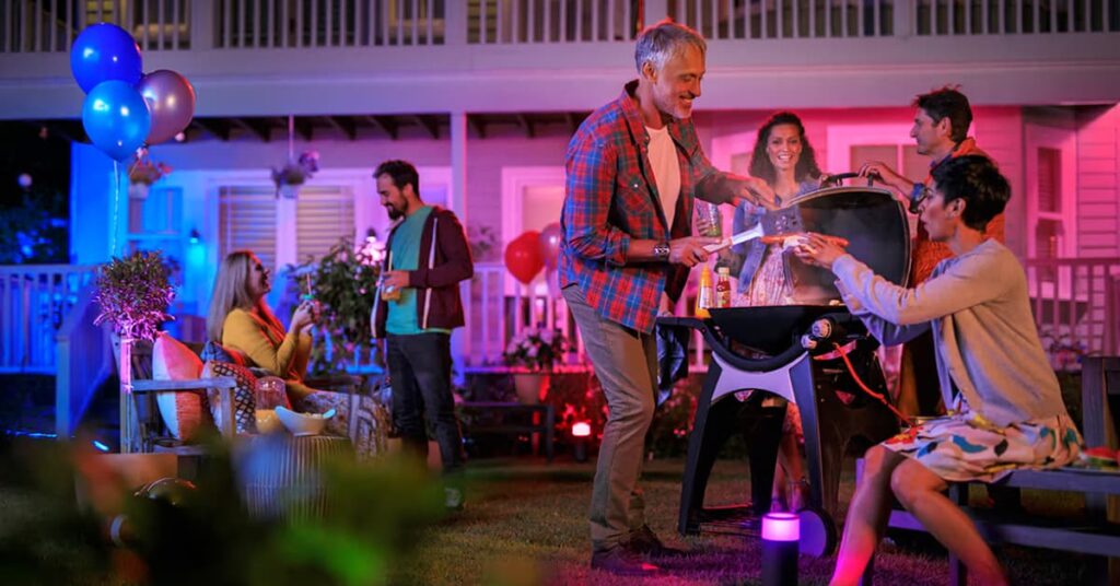 Outdoor party at night with a man grilling and balloons around the yard with pink and blue lighting