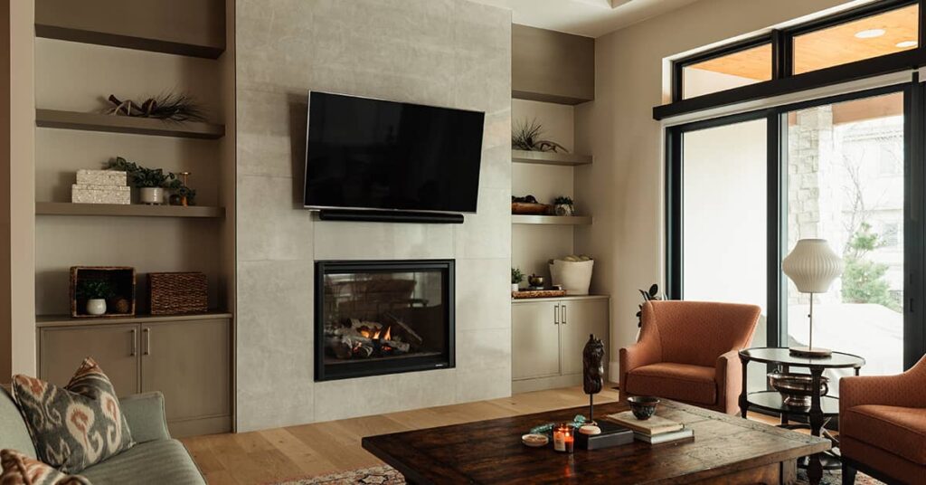 Living area having light colored stone walls with a couch and chairs around a coffee table with a flat screen TV on the wall above a fireplace between two shelving units